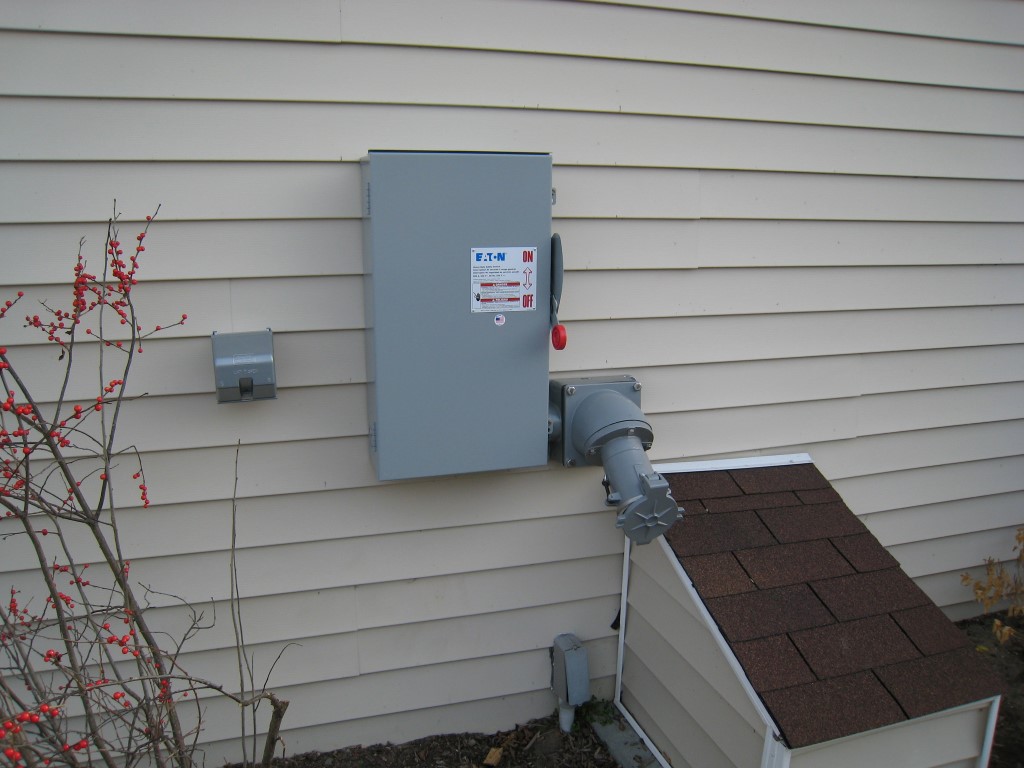 A grey transfer switch and generator inlet mounted on the beige siding of a house, with snow on the ground